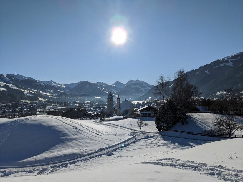 Winterlandschaft bei Achrain Verschneite Hügellandschaft mit Blick auf ein Dorf mit Kirchturm und umliegende Berge unter blauem Himmel.