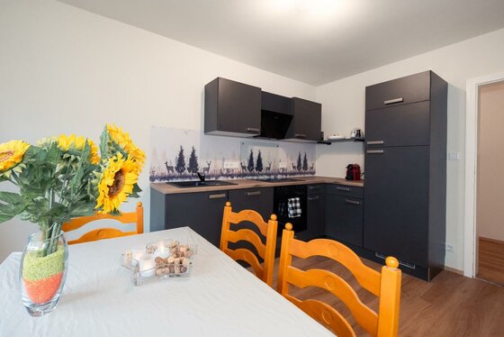 Kitzbühel_DeNi Apt_Küche Large Eine moderne Küche mit grauen Möbeln und einem gemütlichen Esstisch. Auf dem Tisch stehen Sonnenblumen und ein dekoratives Glas mit bunten Elementen.