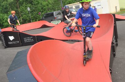 Ein Junge auf einem Roller und zwei Radfahrer befahren einen roten Pumptrack.