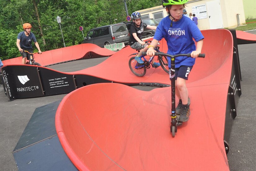 Funpark Pumptrack Ein Junge auf einem Roller und zwei Radfahrer befahren einen roten Pumptrack.