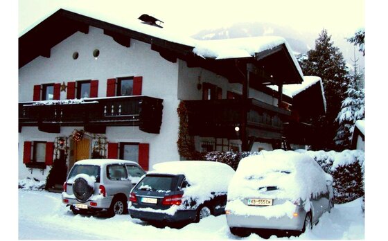 Ein schneebedecktes alpines Haus mit roten Fensterläden und drei parkenden Autos im Vordergrund.