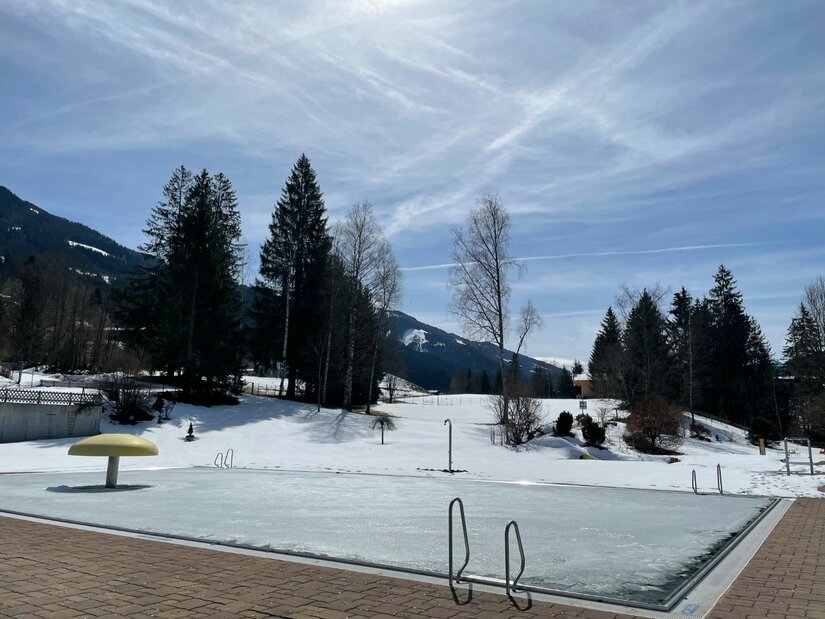 Ein zugefrorenes Freibadbecken in einer verschneiten Berglandschaft mit Nadelbäumen unter blauem Himmel.
