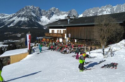 Going_Hollenauer Kreuz_Winter_2022_1 Eine alpine Skisportlandschaft mit schneebedeckten Bergen und einem Holzhaus im Hintergrund. Mehrere Skifahrer und Snowboarder genießen die Piste.