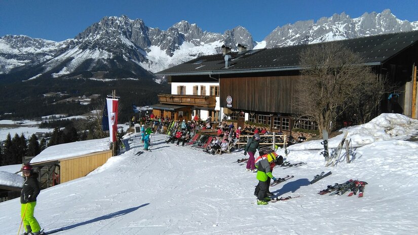 Eine alpine Skisportlandschaft mit schneebedeckten Bergen und einem Holzhaus im Hintergrund. Mehrere Skifahrer und Snowboarder genießen die Piste.