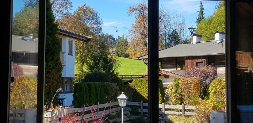 Blick auf die Gondel Blick durch ein Fenster auf alpine Häuser, Gärten und eine Seilbahngondel vor bewaldeten Hügeln.