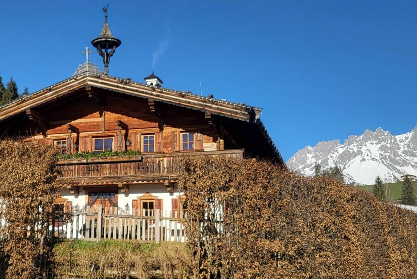 Chalet Nestwärme am Wilden Kaiser Traditionelles Holzhaus mit Balkon vor schneebedecktem Gebirgsmassiv unter blauem Himmel.