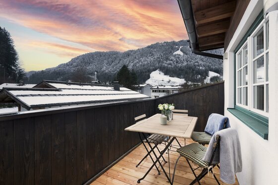 Holzbalkon mit Sitzgruppe vor verschneiten Bergen und Abendhimmel.