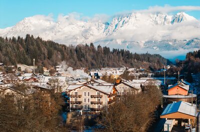 Winteransicht Kitzbühel mit Wildem Kaiser Blick über verschneite Hausdächer in Kitzbühel auf das schneebedeckte Massiv des Wilden Kaisers unter blauem Himmel.