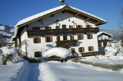Verschneites, weißes Bauernhaus mit Holzbalkonen und braunen Fensterläden unter blauem Himmel.