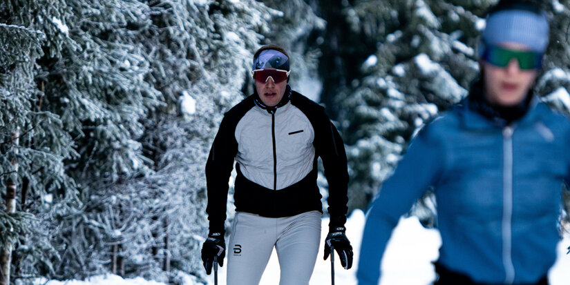 During a cross-country ski race in Kitzbühel, two cross-country skiers glide through a snow-covered forest of tall fir trees.