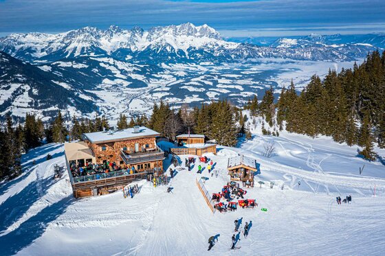Berghütte aus Holz im Schnee mit Skifahrern auf der Terrasse vor einem Gebirgspanorama.