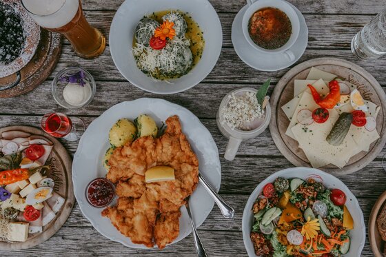 Verschiedene Speisen wie Schnitzel, Knödel, Salat und Brotzeitplatten auf einem Holztisch aus der Vogelperspektive.