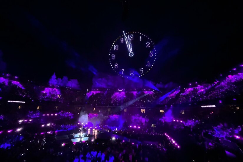 Schlagerbooom Open Air Stadion-Event Blick in ein Stadion bei Nacht mit lila Beleuchtung und einer leuchtenden Uhr am Nachthimmel über der Bühne.