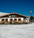 Berghütte aus Holz und Stein mit Terrasse im verschneiten Skigebiet unter blauem Himmel.