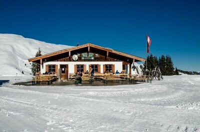 Jausenstation Gauxjoch im Winter Berghütte aus Holz und Stein mit Terrasse im verschneiten Skigebiet unter blauem Himmel.