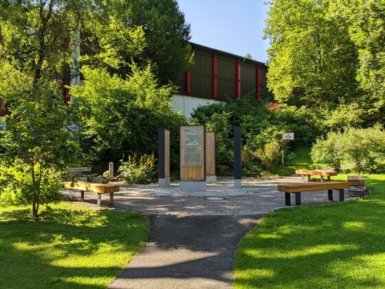 Gepflasterter Platz mit hölzernen Infostelen und Sitzbänken inmitten von Bäumen und Rasenflächen.