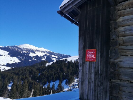 Bruggeralm im Winter Holzhütte mit rotem Schild vor verschneiter Berglandschaft und Nadelwald unter blauem Himmel.