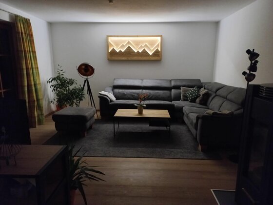 Wohnbereich im Hahnenkammblick Wohnzimmer mit grauem Ecksofa, Holztisch und beleuchtetem Wandbild in Bergform.