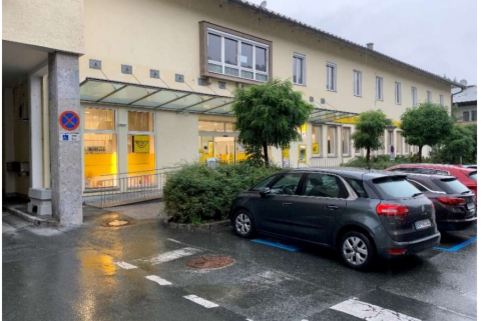 Ein ruhiger Parkplatz mit mehreren Autos vor einem modernen Gebäude. Die Fenster sind beleuchtet und es gibt grüne Pflanzen in der Umgebung.