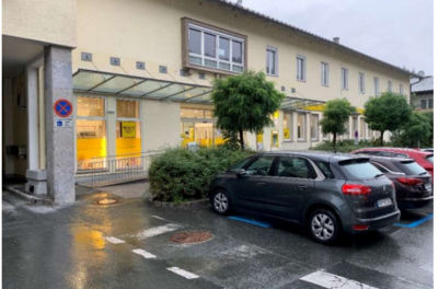Behindertenparkplatz - Josef-Pirchl-Straße 11 Ein ruhiger Parkplatz mit mehreren Autos vor einem modernen Gebäude. Die Fenster sind beleuchtet und es gibt grüne Pflanzen in der Umgebung.
