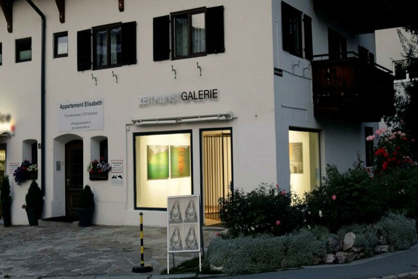 Galerie Zeitkunst Ein Gebäude mit der Aufschrift „Galerie“ auf der Vorderseite. Es befindet sich in einer malerischen Umgebung mit Blumen und einem klaren Himmel.