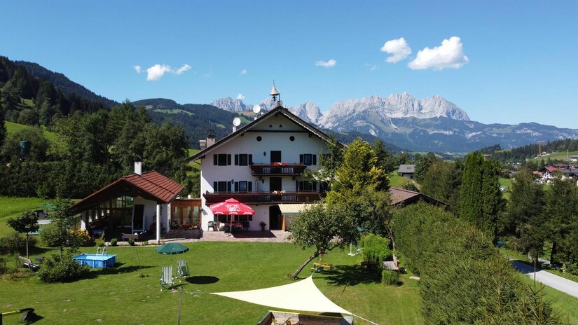 Haus im Sommer Alpines Gästehaus mit Garten und Pool vor einem Gebirgsmassiv unter blauem Himmel.
