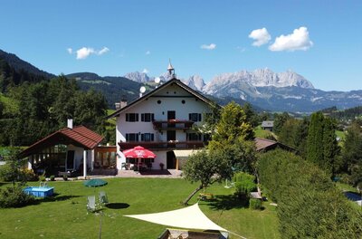 Haus im Sommer Alpines Gästehaus mit Garten und Pool vor einem Gebirgsmassiv unter blauem Himmel.