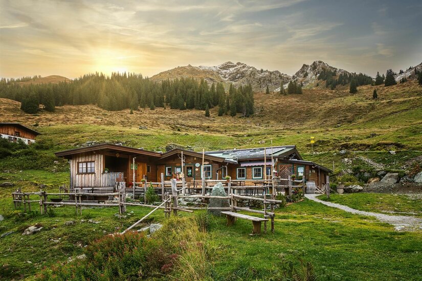 Rustikale Holzhütte mit Terrasse vor bewaldeten Hängen und felsigen Berggipfeln bei tiefstehender Sonne.