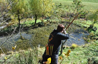Person mit Langbogen zielt von einer Anhöhe über einen Teich auf ein Ziel im Wald.