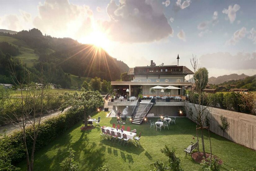 Auwirt im Sommer Außenansicht eines Gasthofs mit Gartenrestaurant, Terrasse und angrenzender Wiese vor bewaldeten Bergen.