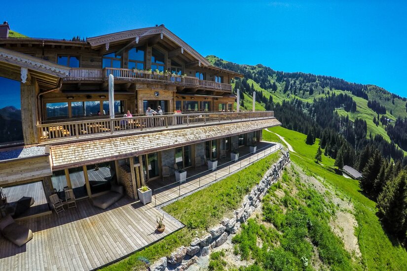 Große Berghütte aus Holz mit Sonnenterrasse am grünen Hang vor bewaldeten Bergen unter blauem Himmel.