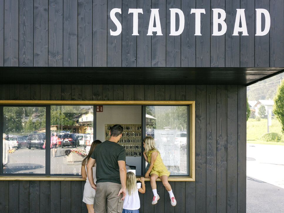 Eine Familie steht vor dem Stadtbad in Kitzbühel, während ein Kind auf die Fensterbank klettert und die anderen zuschauen.
