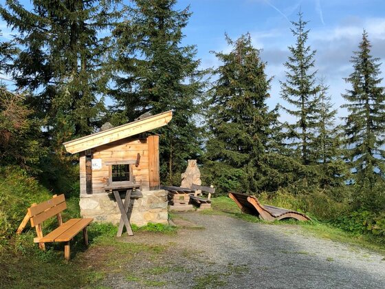 Kleine Holzhütte mit Pultdach, Sitzbänken und einer hölzernen Liege an einem Schotterweg vor Nadelbäumen.