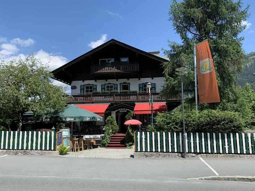 Villa Licht Außenansicht Alpines Gebäude mit Holzbalkonen, grün-weißem Zaun und Außengastronomie unter Sonnenschirmen.