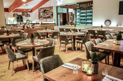 Moderner Restaurantraum mit Holztischen, grauen Polsterstühlen und einem großen Weinregal im Hintergrund.
