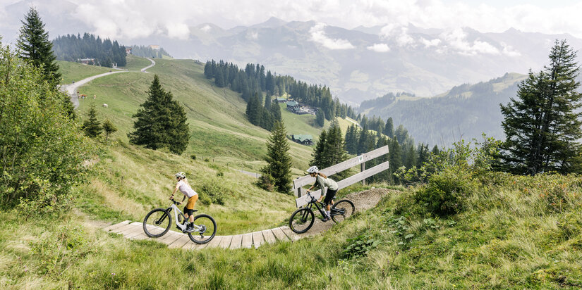 On the 360 Flow Trails in Kitzbühel, two cyclists ride along a winding wooden path through a green mountain landscape dotted with forested hills.