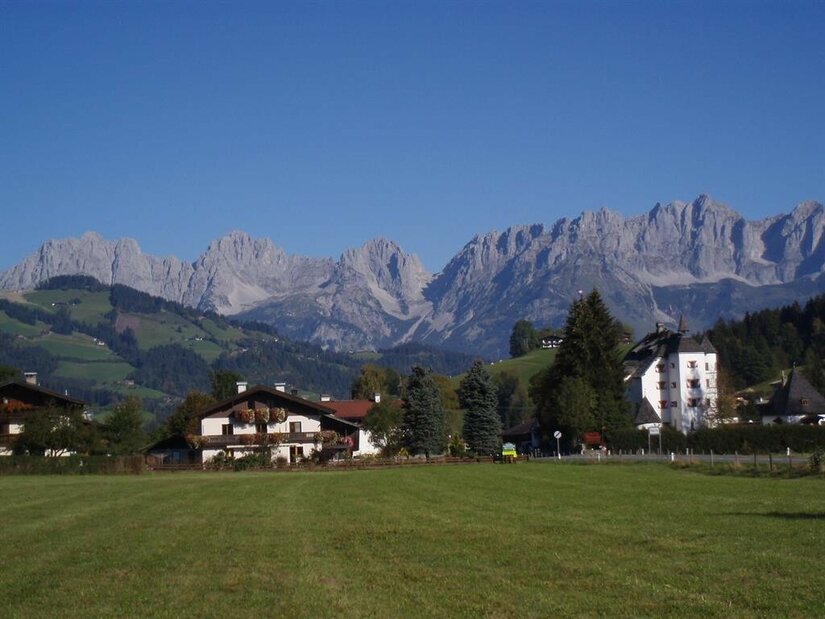 Haus Zierl vor Kaisergebirge Blick über eine grüne Wiese auf das Haus Zierl und ein weißes Schloss vor dem Gebirgsmassiv Wilder Kaiser.