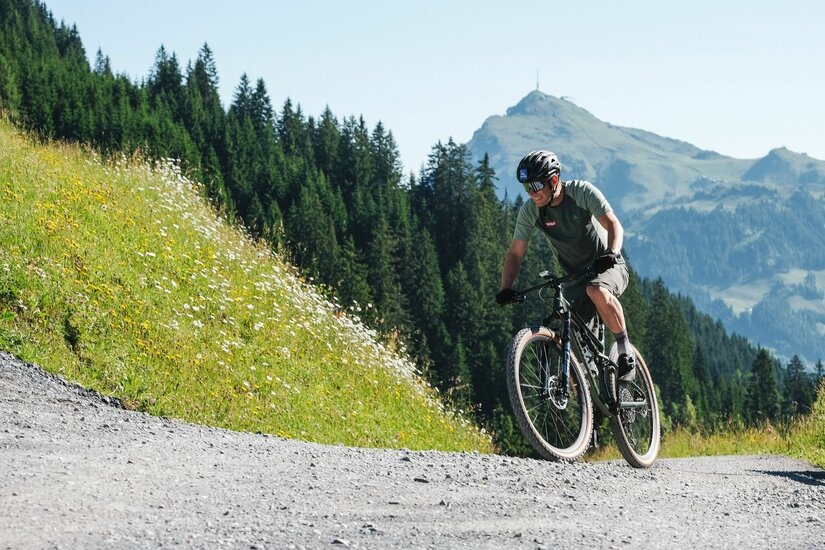 Mountainbiken in Kitzbühel Ein Mountainbiker fährt auf einem Schotterweg vor bewaldeten Bergen und einem Gipfel.