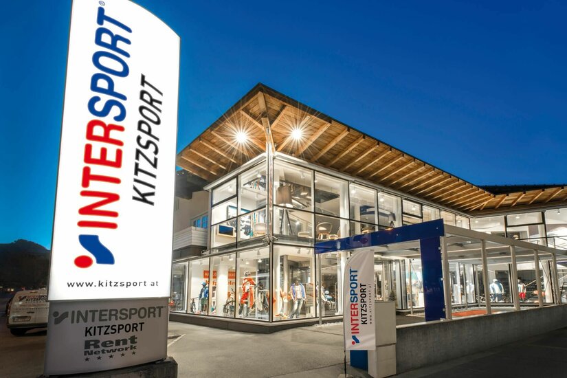 Kitzsport Intersport Filiale Beleuchtetes Sportgeschäft Intersport Kitzsport bei Dämmerung mit großer Werbesäule im Vordergrund.