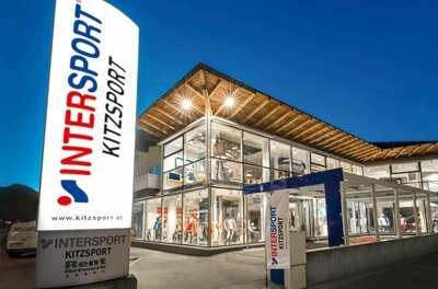 Kitzsport Intersport Filiale Beleuchtetes Sportgeschäft Intersport Kitzsport bei Dämmerung mit großer Werbesäule im Vordergrund.