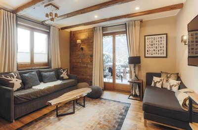 Wohnzimmer Appartement Streifblick Wohnzimmer mit zwei grauen Sofas, Holztisch, Deckenbalken und Balkontür mit Blick auf verschneite Berge.