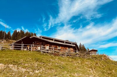 Hütte am Hollenauer Kreuz Rustikale Holzhütte mit Zaun auf einem Grashang unter blauem Himmel.