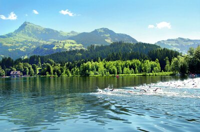 Triathlon Ein ruhiger See umgeben von hohen Bergen und grünen Wäldern. Die Sonne scheint und es gibt einige Menschen, die im Wasser schwimmen.