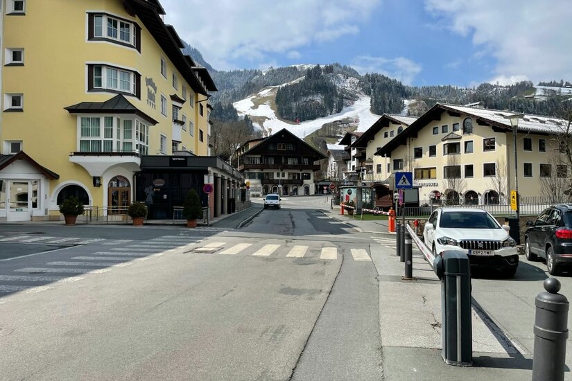 Ortszentrum mit Bergpanorama Blick auf eine asphaltierte Dorfstraße mit mehrstöckigen Gebäuden und schneebedeckten Bergen im Hintergrund.