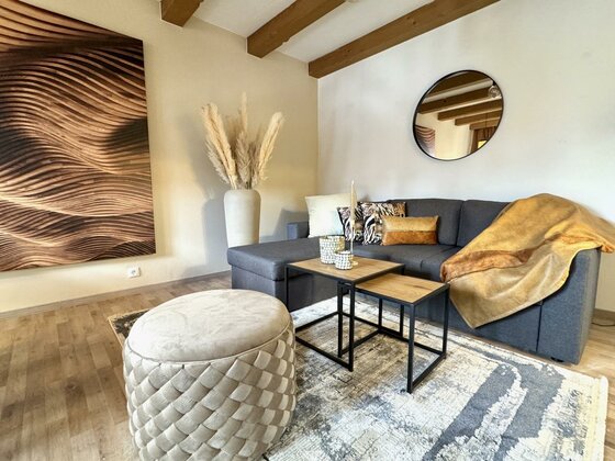 Ein modernes Wohnzimmer mit einer gemütlichen Couch und dekorativen Kissen. Ein stilvoller Tisch und ein runder Pouf ergänzen den Raum, der mit einer Kunstwand und Pflanzen dekoriert ist.