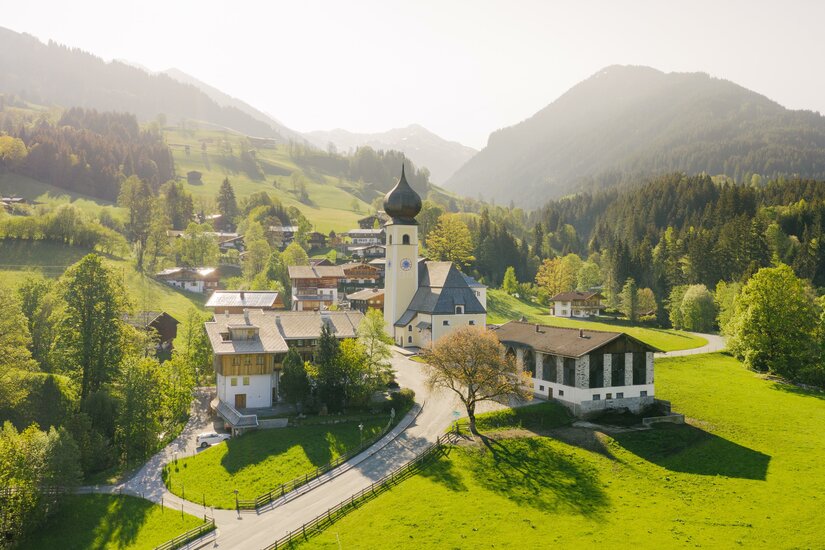 Dorfansicht Aurach Eine malerische Landschaft mit grünen Hügeln und einer kleinen Kirche. Im Hintergrund sind Berge und ein klarer Himmel zu sehen.