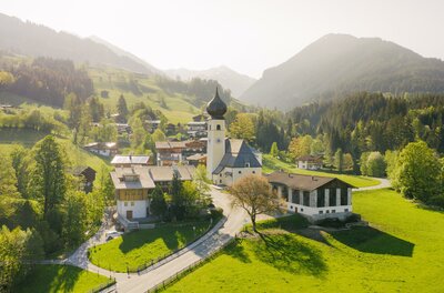 Eine malerische Landschaft mit grünen Hügeln und einer kleinen Kirche. Im Hintergrund sind Berge und ein klarer Himmel zu sehen.
