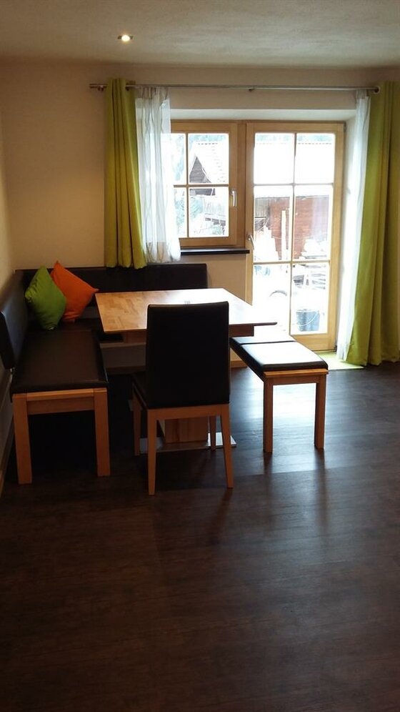 Wohnung EG5 Biohof Foidl Sitzecke mit Holztisch, Eckbank und Stuhl vor einem Fenster mit grünen Vorhängen.