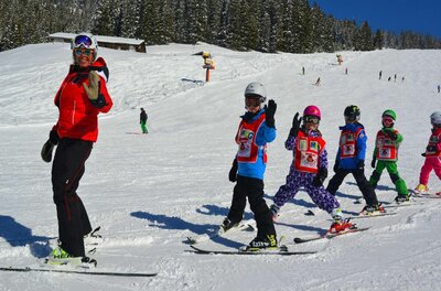 Skilehrerin in roter Jacke mit einer Gruppe von Kindern in bunten Skianzügen auf einer verschneiten Piste.