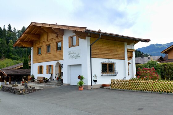 Landhaus Marie Luise Zweistöckiges Landhaus mit weißer Fassade, Holzverkleidung im Obergeschoss und flachem Satteldach vor Bergkulisse.